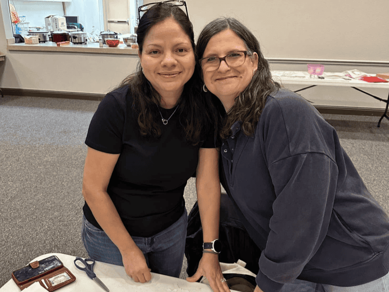 Two people smiling at a table in a room with food and craft supplies. They appear to be enjoying their time together.