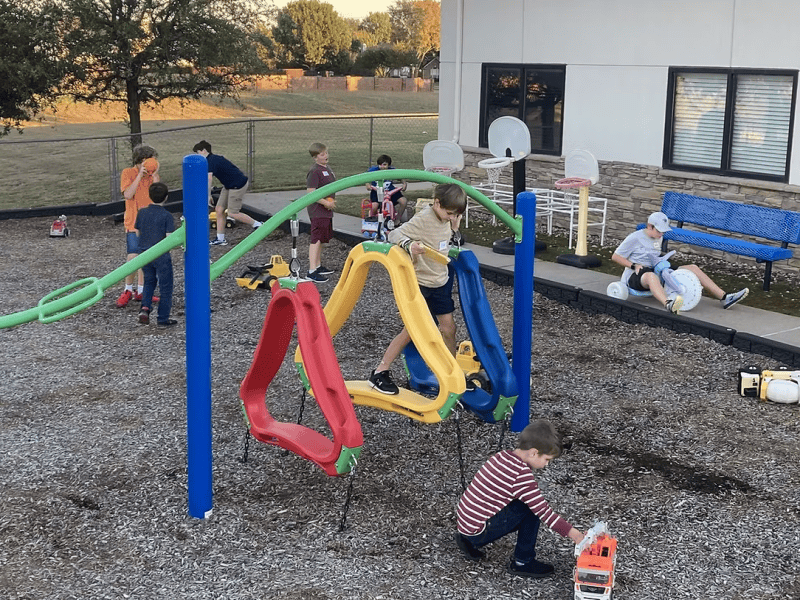 Children play in a colorful playground, climbing and riding toys. A person sits nearby. Trees and a building form the background.